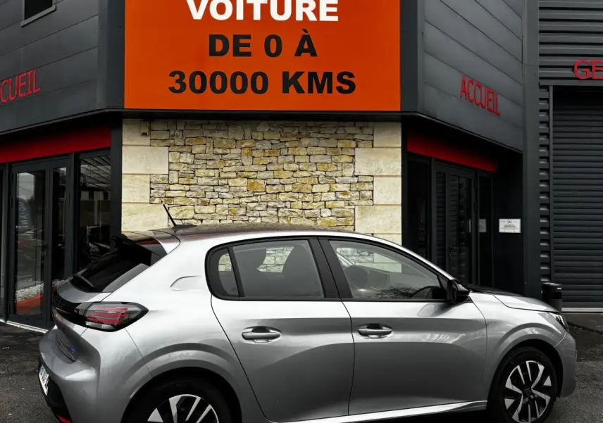 Vue latérale droite d'une Peugeot 208 PureTech 100 S&S Active gris clair stationnée devant un bâtiment avec panneau orange.