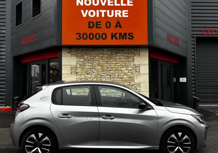 Profil droit d'une Peugeot 208 PureTech 100 S&S Active gris clair garée devant un bâtiment avec panneau orange.