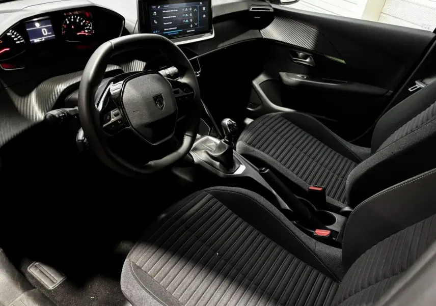 Vue intérieure côté conducteur de la Peugeot 208 PureTech 100 S&S Active 2024 avec tableau de bord numérique et sièges noirs.