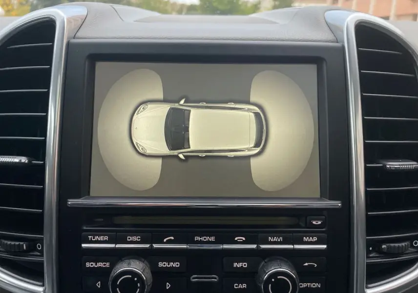 Écran central du Porsche Cayenne S Hybrid 2010 montrant une vue aérienne du véhicule gris avec capteurs avant et arrière activés.