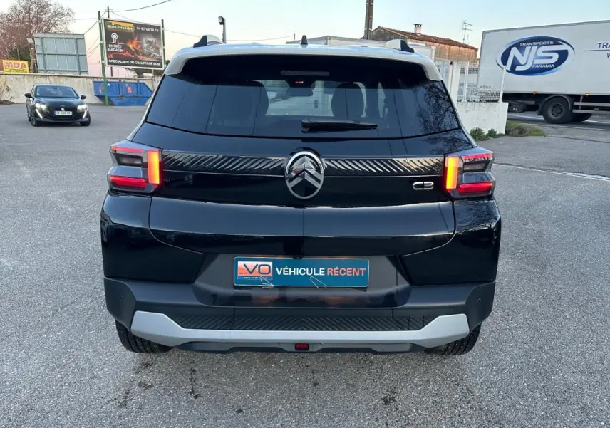 Vue arrière d'une Citroën C3 noire 2025 avec feux allumés et barres de toit visibles sur un parking.