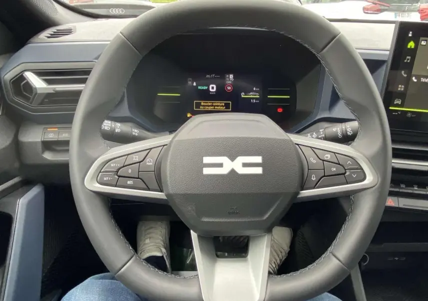 Vue intérieure centrée sur le volant noir du Dacia Duster 1.6 Hybrid 2025 avec tableau de bord digital allumé.