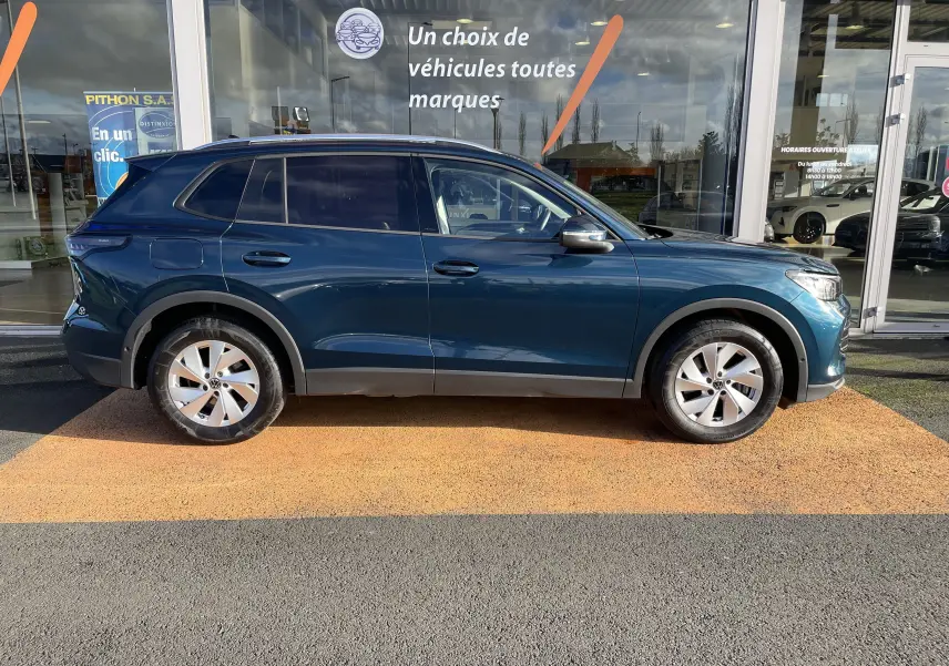 Vue de profil côté gauche d’un Volkswagen Tiguan 2.0 TDI 150 DSG Life Plus 2024 en bleu Nightshade, stationné devant une vitrine.