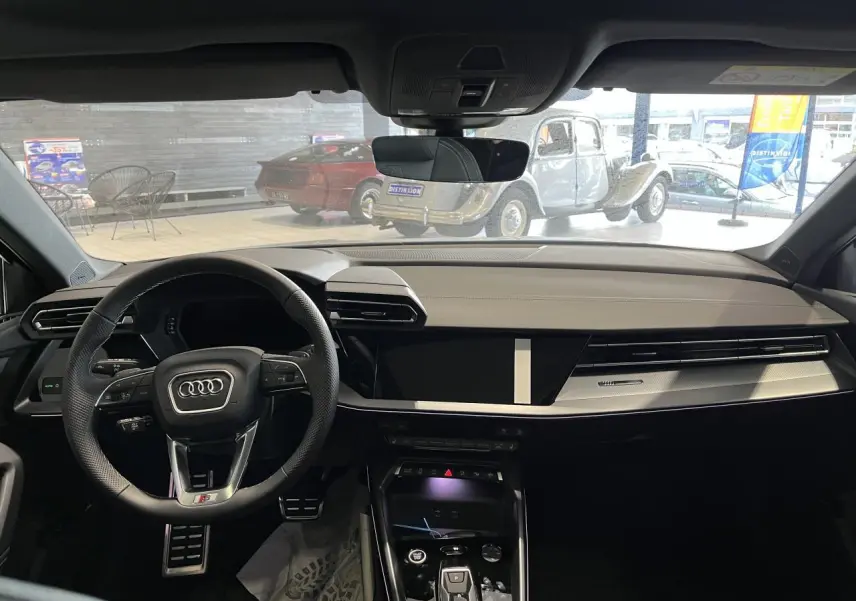Vue intérieure avant de l'Audi A3 Sportback 2025 gris, tableau de bord moderne avec volant sport multifonctions et écran tactile central.