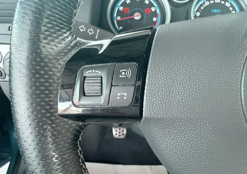 Gros plan sur le volant noir perforé de l’Opel Astra OPC 2008 avec commandes intégrées et compteurs bleus en arrière-plan.