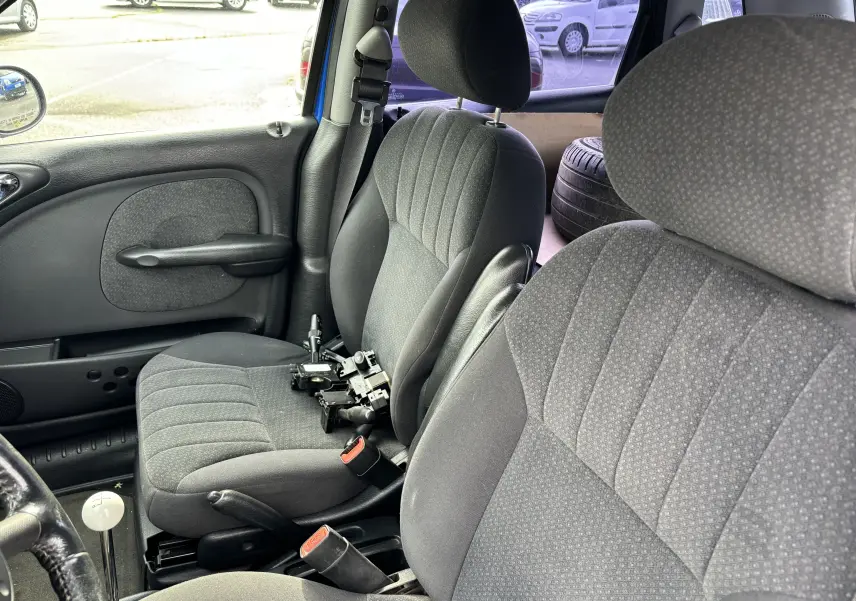 Vue intérieure côté gauche du Chrysler PT Cruiser bleu de 2004, montrant les sièges avant en tissu gris et une pièce mécanique posée.