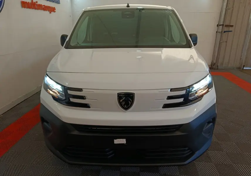 Vue de face du Peugeot Partner blanc 2025 avec phares allumés et calandre noire distinctive dans un garage.