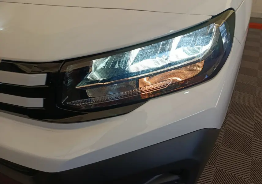 Gros plan sur le phare avant droit du Peugeot Partner blanc 2025, avec calandre noire et détails lumineux distinctifs.