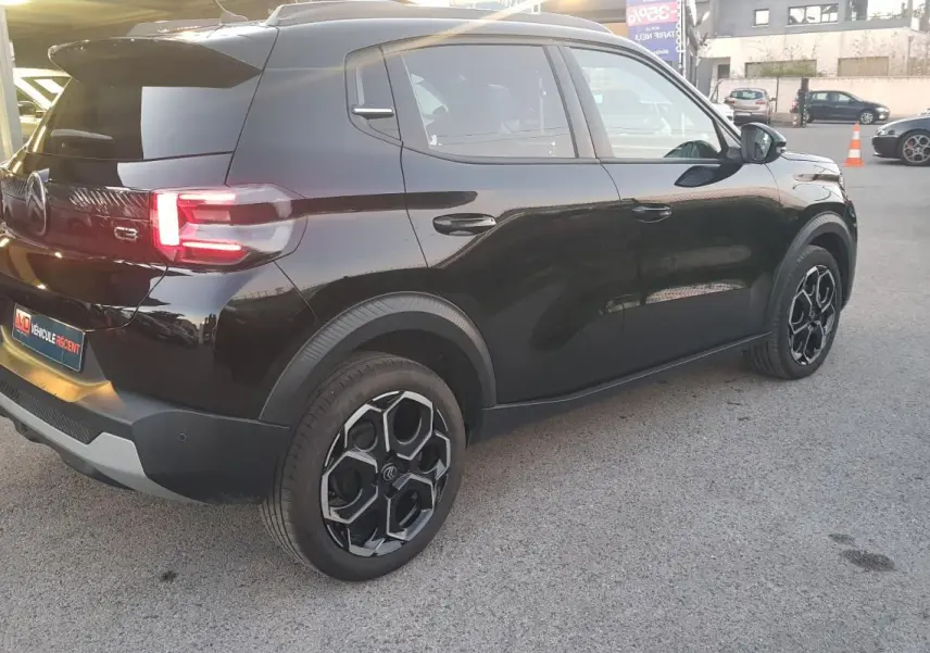 Vue 3/4 arrière droite d'une Citroën C3 noire 2025 avec jantes alliage noires et barres de toit visibles.