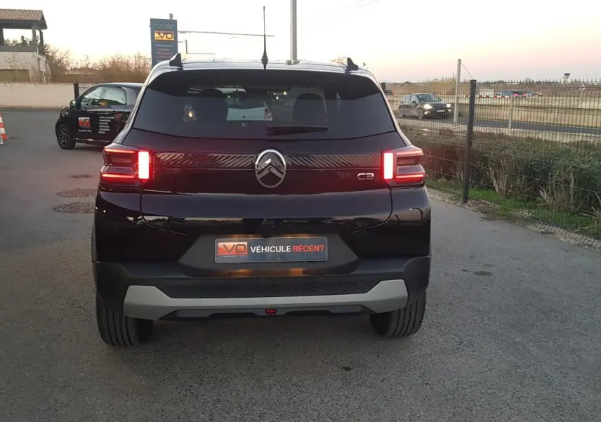 Vue arrière d'une Citroën C3 noire 2025 avec feux allumés et barres de toit visibles sur un parking extérieur.