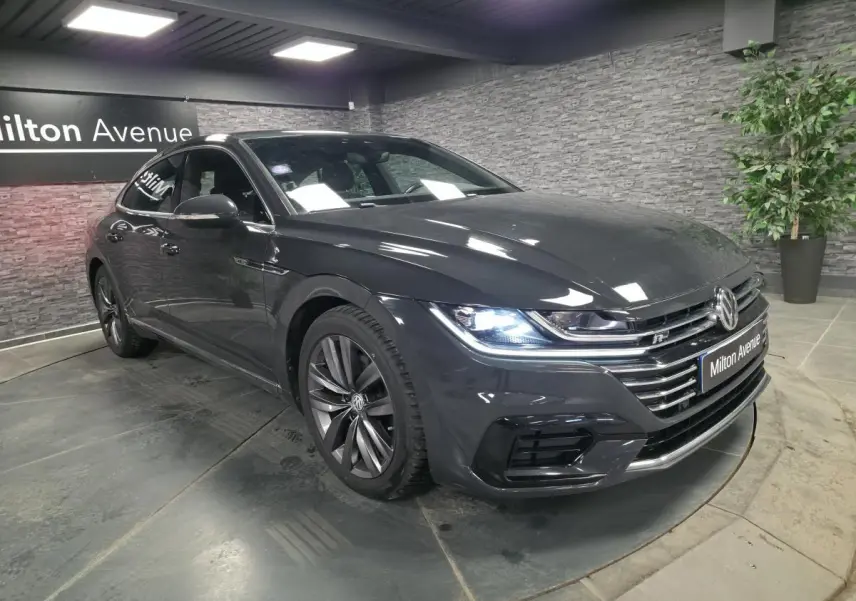 Volkswagen Arteon gris 3/4 avant droit en intérieur, avec phares LED allumés et badge R-Line visible.