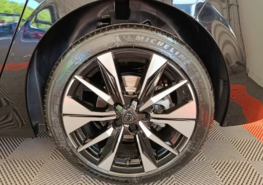 Gros plan sur la roue avant droite noire d'une Peugeot 308 2026 avec jante alliage bicolore et logo lion central.