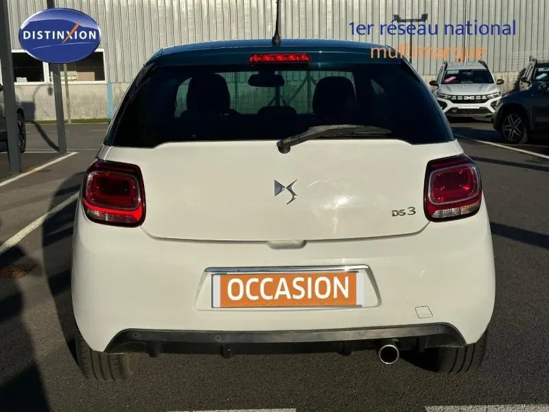 Vue arrière d'une DS3 blanche avec toit noir et feux arrière rouges, stationnée en extérieur sur parking.
