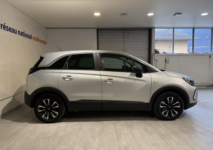 Vue de profil côté droit d’un Opel Crossland gris 2023 avec jantes noires et toit blanc en intérieur showroom.
