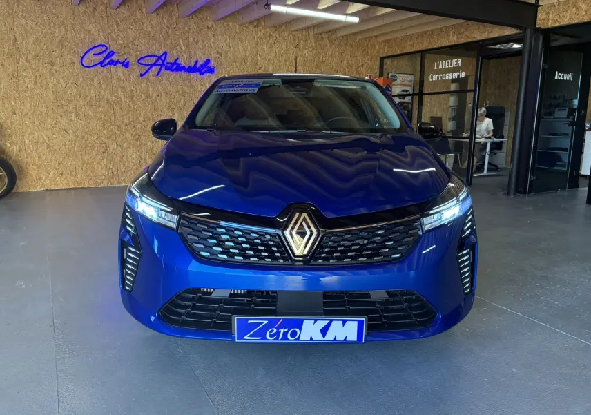 Vue frontale d'une Renault Clio TCe 90ch X-Tronic Evolution bleu iron avec calandre noire et phares allumés en intérieur showroom.