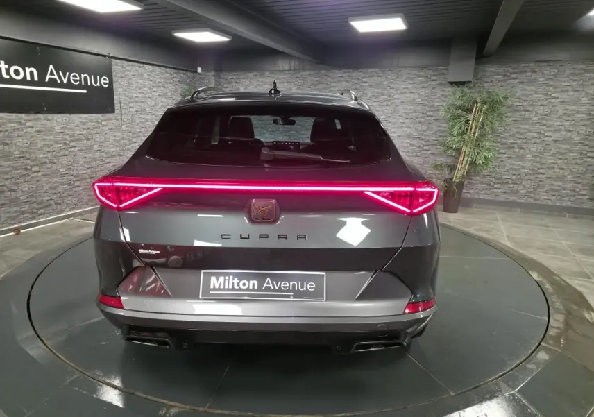 Vue arrière d'une CUPRA Formentor gris caïman avec bandeau lumineux LED et double sortie d'échappement noire dans un showroom.