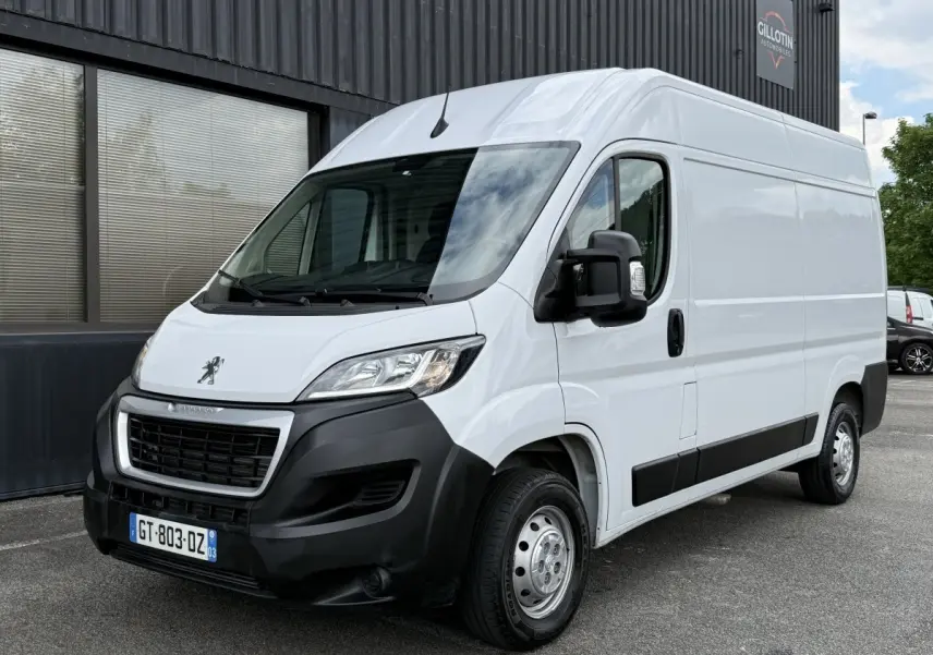 Vue 3/4 avant droit d’un Peugeot Boxer Fourgon blanc avec pare-chocs noir et rétroviseurs noirs, stationné devant un bâtiment.