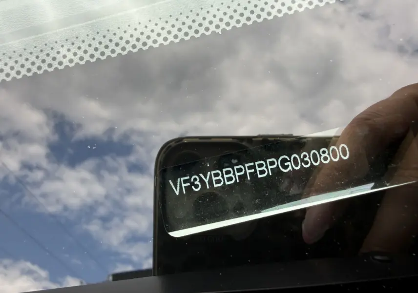 Vue rapprochée du pare-brise avec numéro VIN visible, reflet du ciel et main tenant un smartphone.