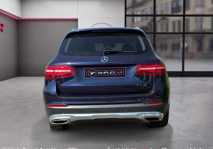 Vue arrière d'un Mercedes GLC 350 E HYBRID FASCINATION 4MATIC bleu avec feux allumés et double sortie d'échappement chromée.
