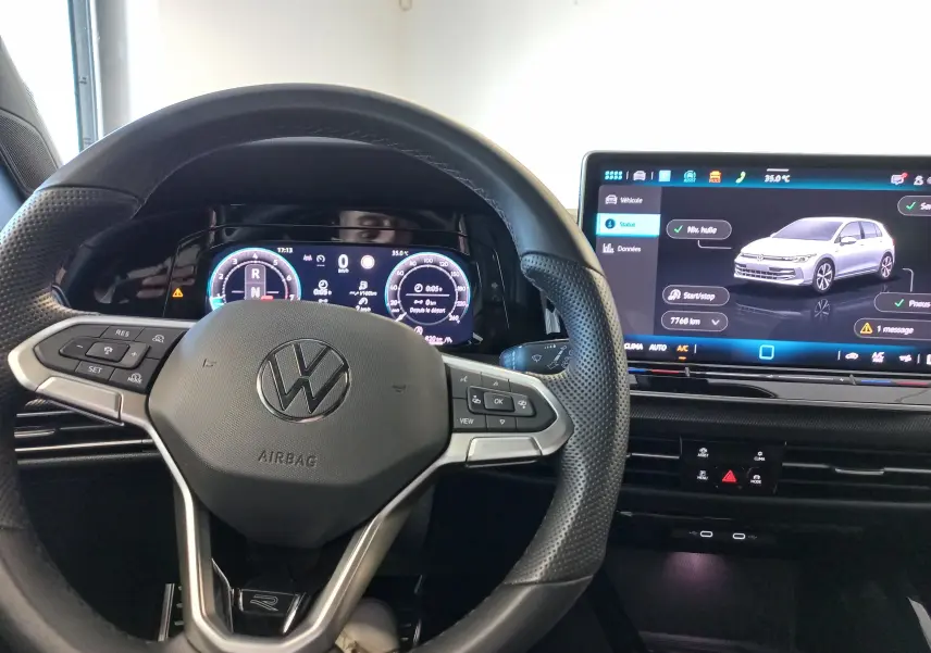 Vue intérieure centrée sur le volant Volkswagen et l'écran digital affichant une Golf gris dauphin en 3D.