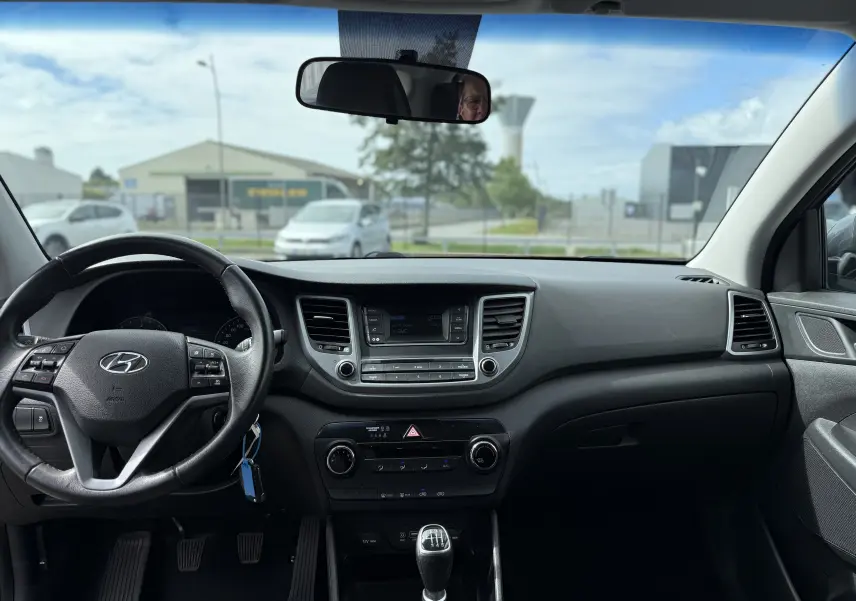 Vue intérieure du tableau de bord et volant d'un Hyundai Tucson gris 2015 avec boîte manuelle, climatisation et écran central.