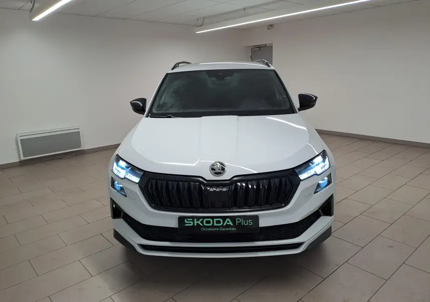 Vue frontale d’un Skoda Karoq blanc lune 2022 avec phares LED allumés et calandre noire Sportline dans un intérieur neutre.