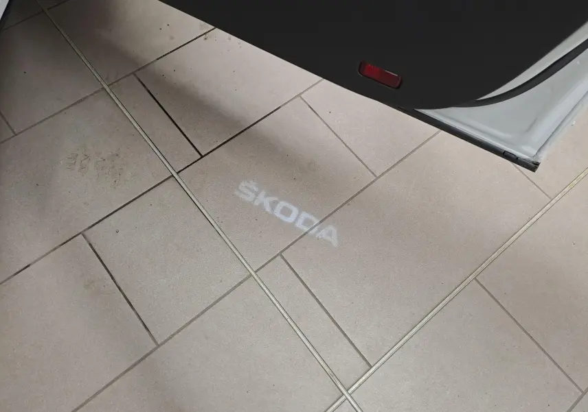 Projection lumineuse du logo SKODA au sol depuis la porte ouverte d'un SKODA KAROQ blanc lune 2022 côté droit.
