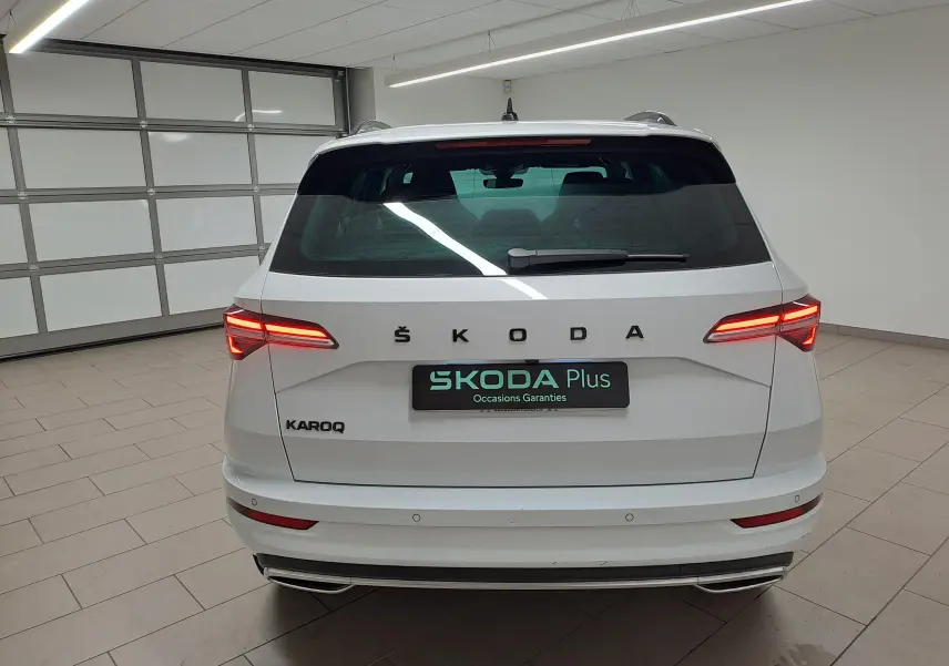 Vue arrière d'un Skoda Karoq blanc Lune 2022 avec feux allumés et logo ŠKODA bien visible sur un fond intérieur neutre.