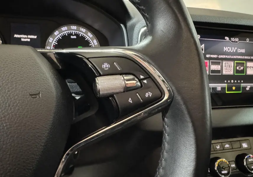 Gros plan sur la partie gauche du volant cuir noir du Skoda Karoq blanc, avec commandes multifonctions et écran tactile en arrière-plan.