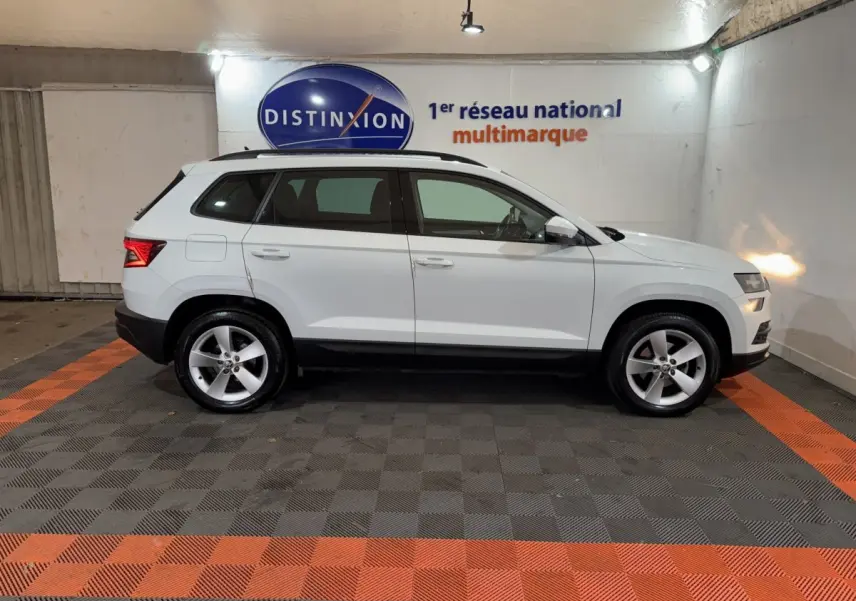 Profil droit d'un SUV SKODA KAROQ blanc 2021, avec jantes alliage et rails de toit noirs, en intérieur showroom.