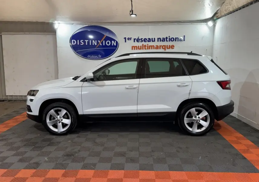 Vue latérale droite d'un SKODA KAROQ blanc 2021 avec barres de toit noires dans un showroom.