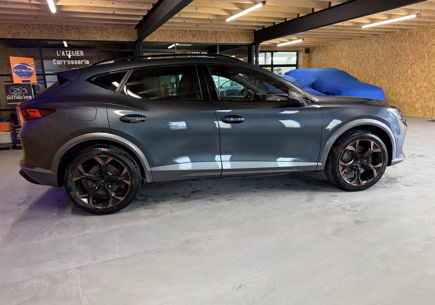 Vue de profil côté gauche d'un CUPRA Formentor gris clair 2023 avec jantes cuivrées et lignes sportives dans un garage.