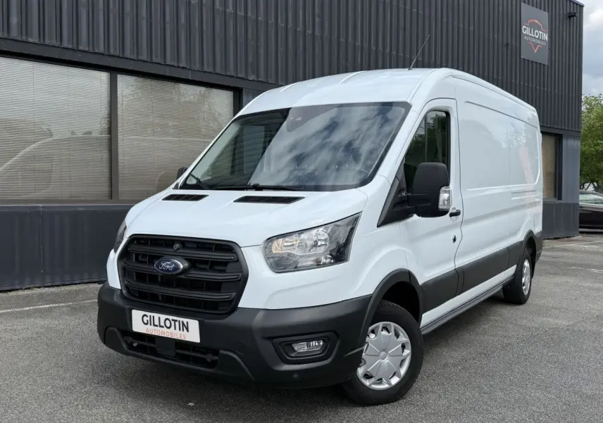 Ford Transit Fourgon blanc vu en 3/4 avant droit, avec calandre noire et feux allumés devant un bâtiment industriel.