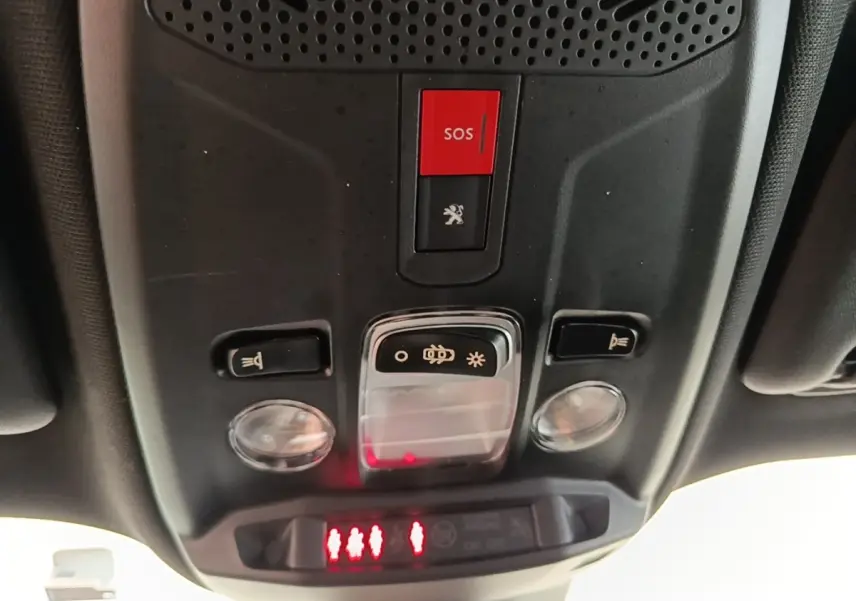 Vue en contre-plongée du panneau de commande de toit intérieur du Peugeot E-2008 électrique gris clair, avec bouton SOS rouge et éclairage.