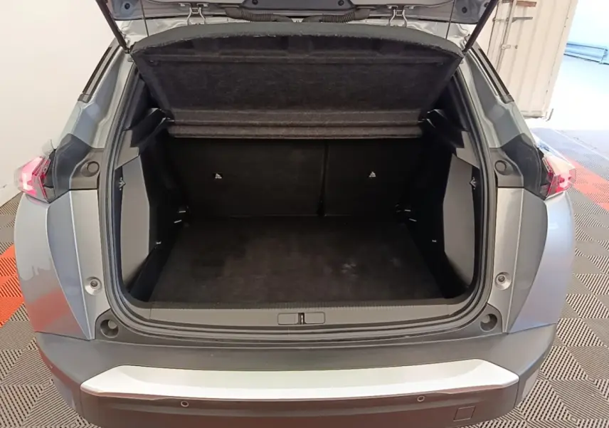 Coffre ouvert vu de l'arrière d'un Peugeot E-2008 électrique gris clair, montrant l'espace de chargement vide.