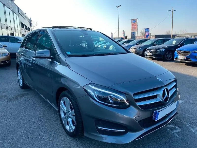 Vue 3/4 avant d'une Mercedes Classe B 180 d gris foncé métallisé, garée en extérieur sur un parking.