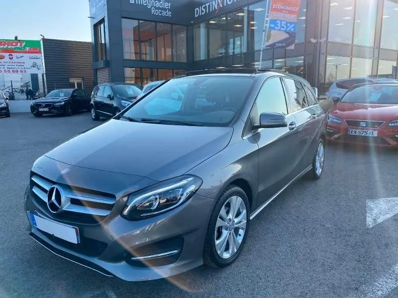 Mercedes Classe B 180 d 2016 gris foncé métal vue 3/4 avant droit sur parking devant un bâtiment commercial.