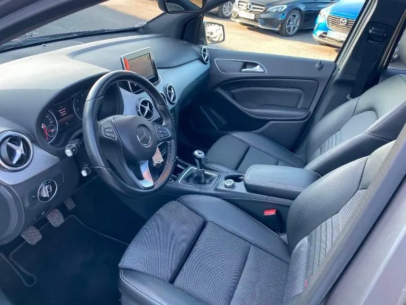 Vue intérieure côté conducteur de la Mercedes Classe B 180 d 2016, avec volant cuir, tableau de bord moderne et boîte manuelle.