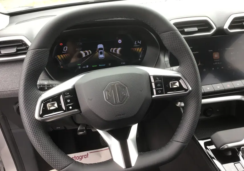 Vue intérieure centrée sur le volant noir perforé du MG ZS 1.5 L Hybrid+ avec tableau de bord numérique et écran tactile.