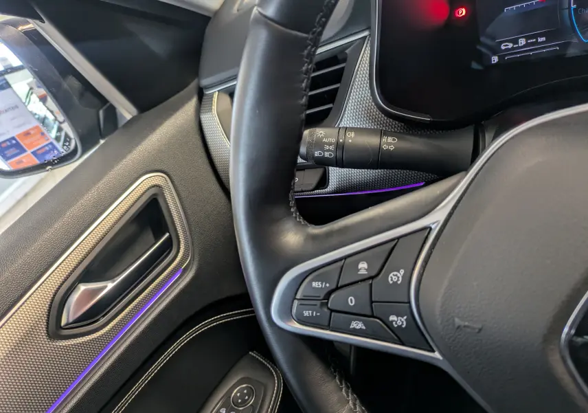 Vue rapprochée de l’intérieur côté gauche du Renault Arkana noir métal, montrant le volant avec commandes et la portière avec éclairage d’ambiance violet.