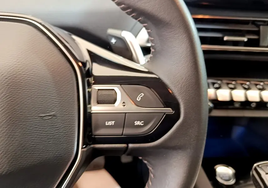 Gros plan sur le volant cuir multifonctions côté gauche du Peugeot 5008 bleu, avec commandes intégrées et tableau de bord en arrière-plan.
