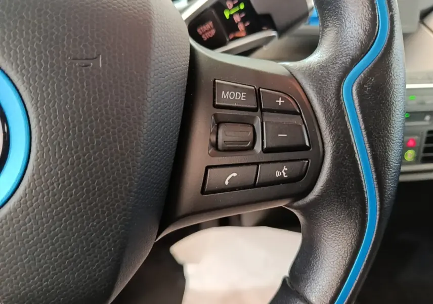 Gros plan sur le volant noir avec surpiqûres bleues de la BMW i3 120 Ah Atelier, montrant les commandes multifonctions.