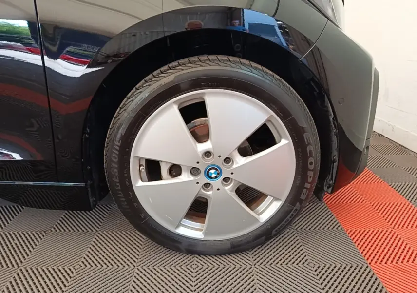 Gros plan sur la roue avant droite noire d’une BMW i3 120 Ah Atelier, avec jante argentée et logo BMW central.