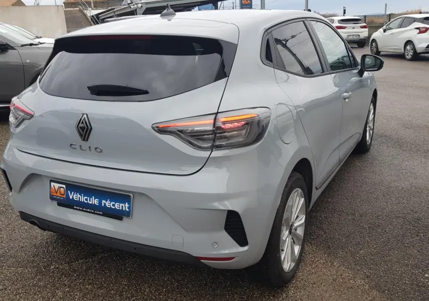 Vue 3/4 arrière droite d'une Renault Clio gris clair avec vitres surteintées et feux arrière allumés.