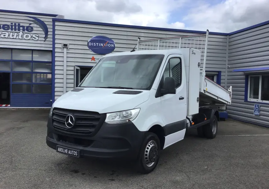Vue 3/4 avant droite d'un Mercedes Sprinter blanc avec benne basculante et coffre latéral, stationné devant un garage.