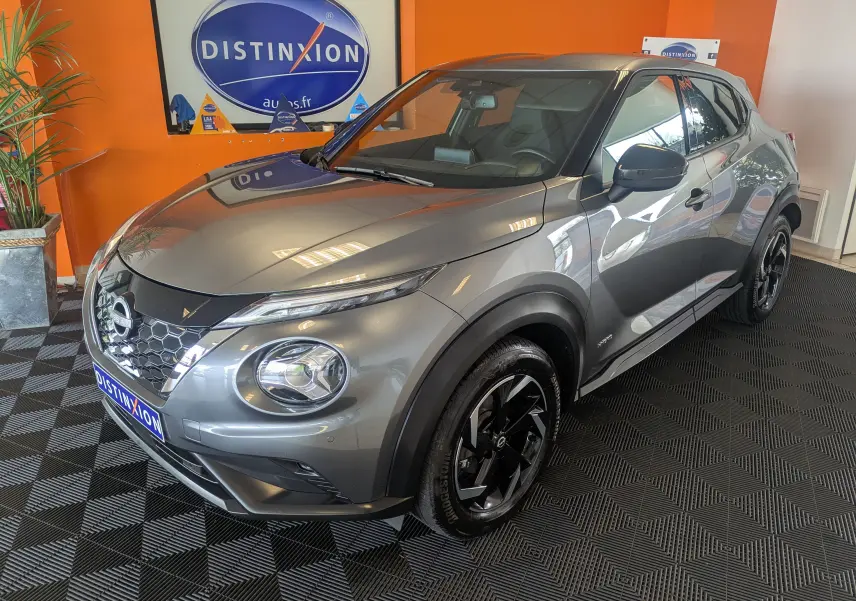 Nissan Juke 1.6 HEV gris squale métal vu en 3/4 avant droit, avec jantes noires et calandre hexagonale distinctive.