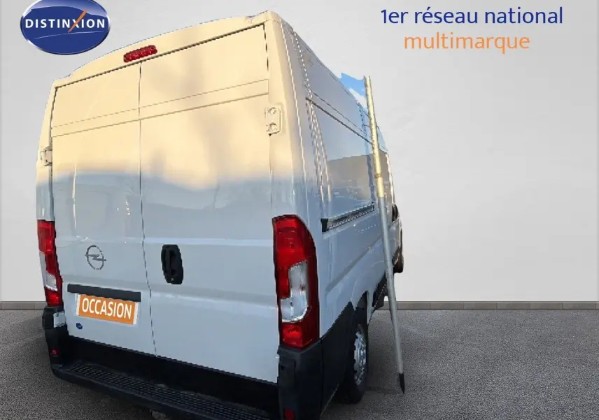 Vue 3/4 arrière droite d’un Opel Movano Fourgon blanc Icy 2023 avec porte arrière fermée et panneau publicitaire à côté.