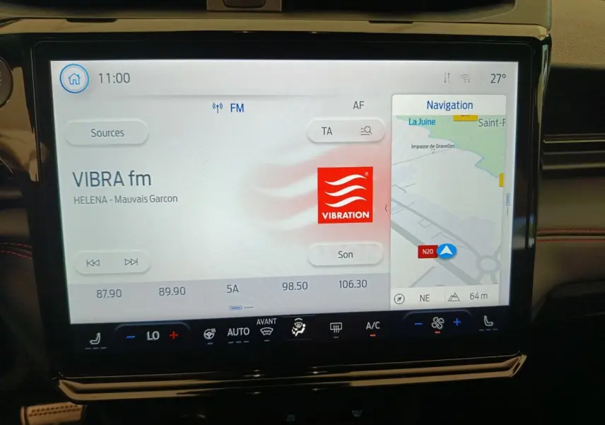 Écran tactile central du Ford Puma 2025 affichant radio et navigation, avec tableau de bord noir et surpiqûres rouges visibles.