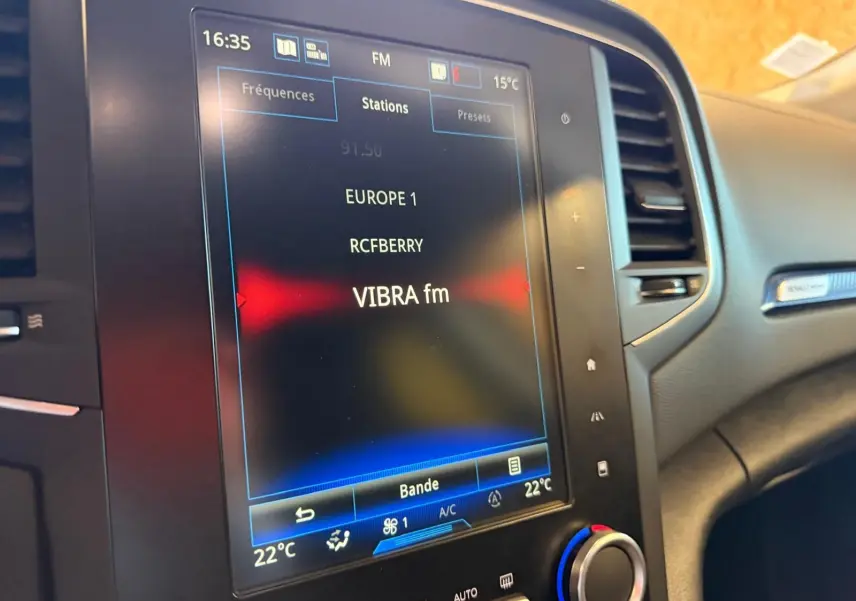Écran tactile central du tableau de bord de la Renault Mégane Break gris, affichant les stations radio FM avec climatisation à 22°C.