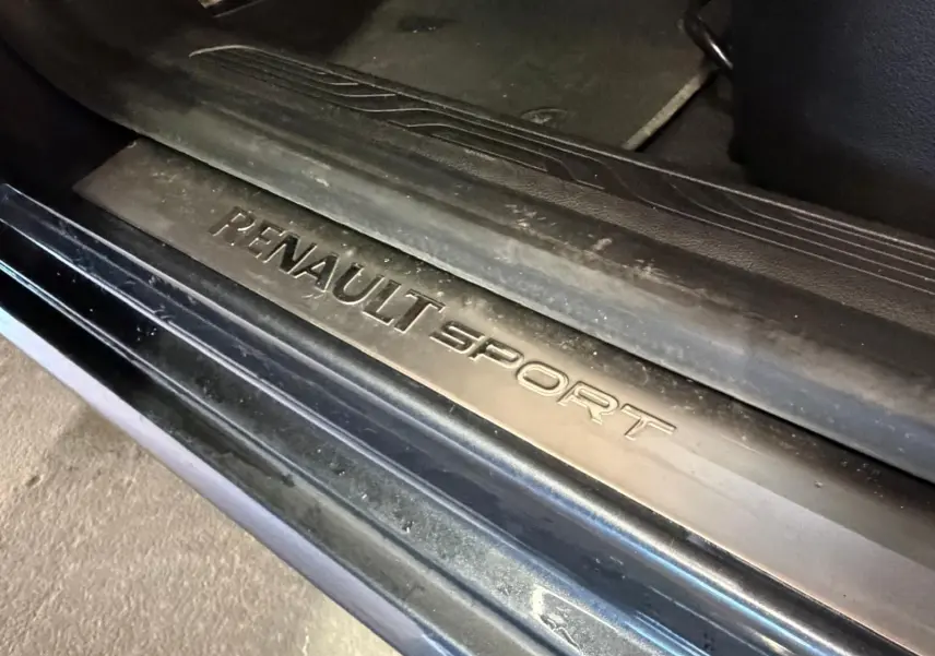 Gros plan sur la plaque de seuil de porte "RENAULT SPORT" noire, intérieur sombre du véhicule visible.